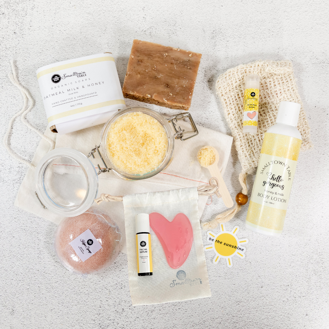 Spa Gift products laid out with a top view. Soap Bar, one packaged one naked, Bath salt jar, facial sponge, facial serum, Gua sha, be the sunshine sticker, bottle of body lotion, lip balm on a soap sack