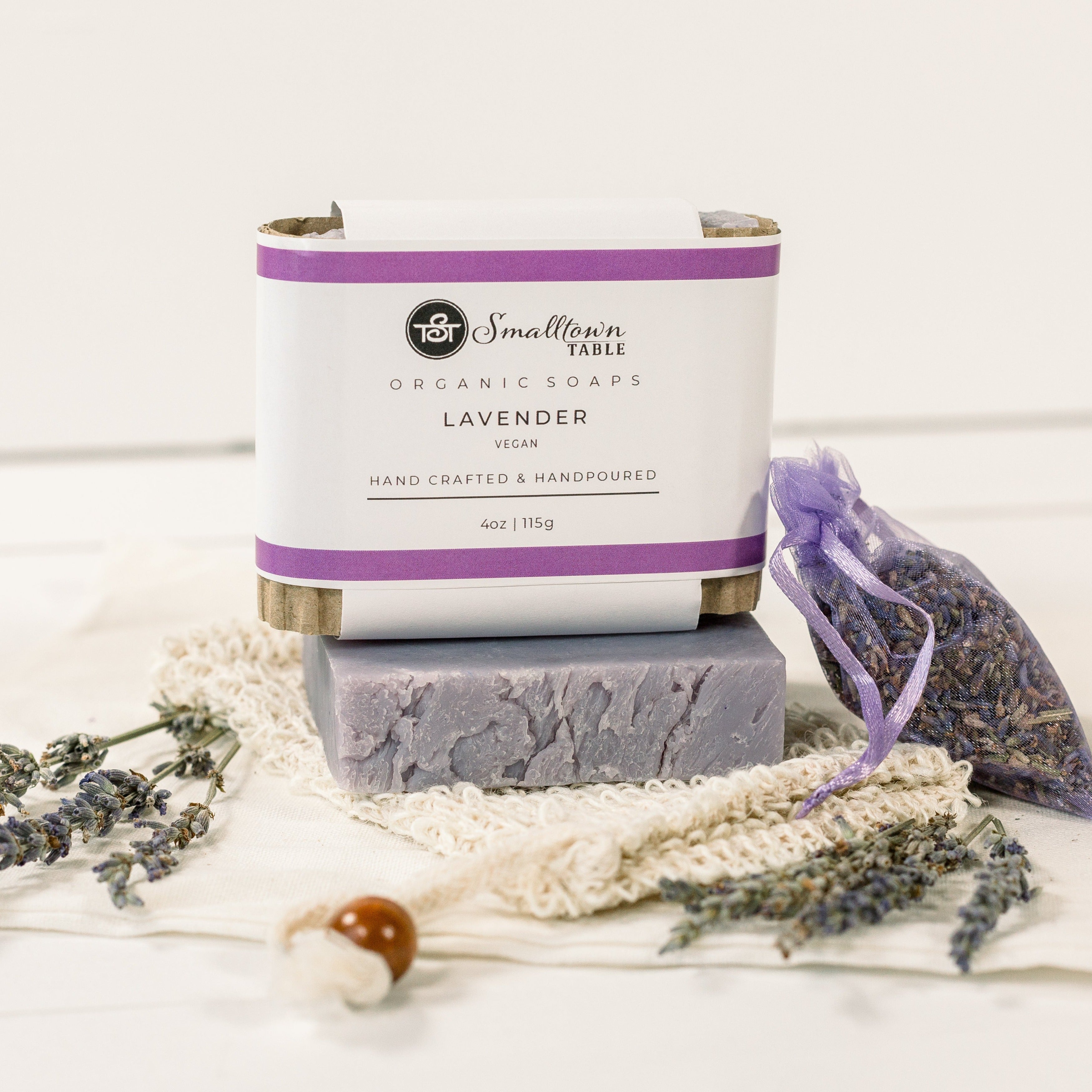 lavender soap wrapped inwhite with purple stripes on top of an unpackaged soap sitting on a soap saver bag with lavender buds in a sack on the right