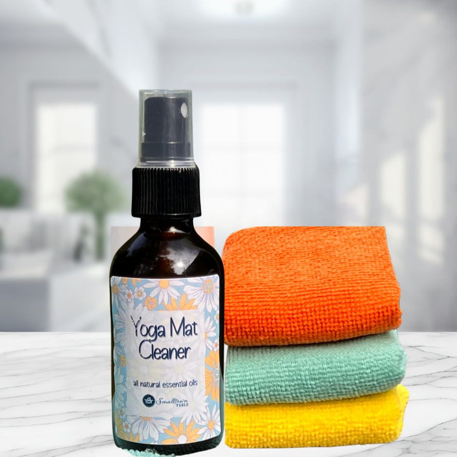 yoga mat cleaner in amber bottle with flowery labels sitting on marble counter with 3 folded small towels 1 orange, 1 turquoise, 1 yellow piled next to it