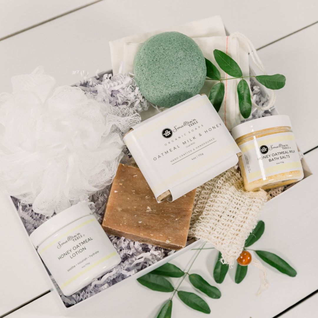 Gift box full of oatmeal milk and honey self-care products. Soap bar on top, ja of lotion, bath salts, soap sack and gift set in white gift box
